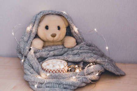 A white teddy bear is wrapped in scarf, next to cup of hot chocolate with marshmallows in the garland lightsの写真素材