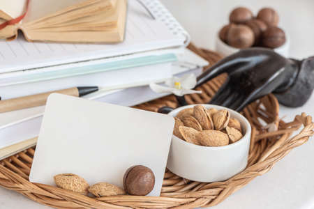 A stack of books and notebooks, a white blank card, a snack of nuts on a table in a wicker tray....
