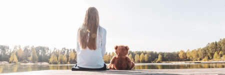 Little girl with stuffed toy teddy bear sitting on wooden pier near calm lakeの写真素材