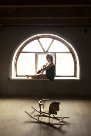 Beautiful curly young girl sits on the window in the attic and looks at the childs wooden horse. A woman Thinks about her childhood and is sadの写真素材
