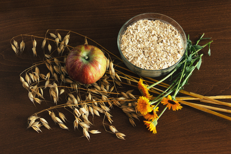 Oat ears stems and oat flakes in a white pot, marigold flowers and apple on dark brown wood table. Top view. oat flakes small size grind. Dietary breakfastの写真素材