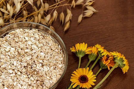 Oat ears stems and oat flakes in a bowl on a dark brown wood background. Top view. oat flakes small size grind. Flower of calendula. Useful fiber-rich product. Dietary breakfast from healthy foodsの写真素材