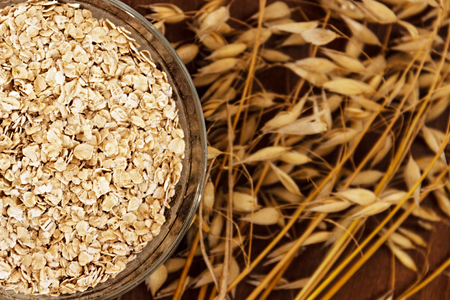 Oat ears stems and oat flakes in a bowl on a dark brown wood background. Top view. oat flakes small size grind. Copy space. Close up. Useful fiber-rich product. Dietary breakfast from healthy foodsの写真素材