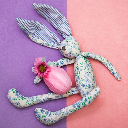 Creative funny bunny from blue, white fabric holds an Easter egg in his hands. egg is wrapped in gift pink paper with mint green ribbon and flower. pink and purple felt background. Consept of Easter.の写真素材