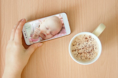 view handheld color video baby monitor. Female hands are holding a smartphone with a baby monitor app. Near hot drinkの写真素材