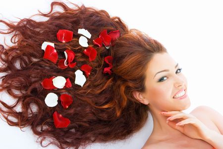 Close-up beautiful luxury fresh bright young lady lying in studio shot with rose petals in her red long hairの写真素材