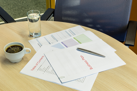 Business plan on a wood desk with coffee, glass of water, an expensive pen and a chair on the background. Top view,の写真素材