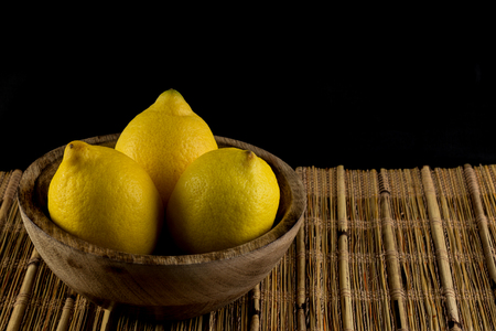 Three lemons in a natural handmade real wood bowl on a bamboo place mat isolated in black backgroundの写真素材