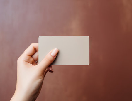 Female hand holding blank business card on brown background. Mockup for branding identity.の素材