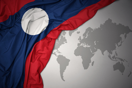 waving colorful national flag of laos on a gray world map background.の写真素材