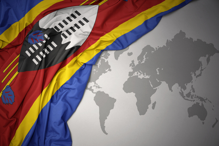 waving colorful national flag of swaziland on a gray world map background.の写真素材
