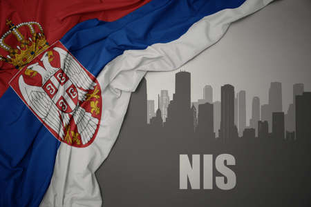 abstract silhouette of the city with text Nis near waving colorful national flag of serbia on a gray...