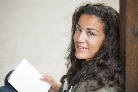 Sitting on street, a young woman (20s) with a book on knees looking at camera. She is relaxed and smiling. Closeup and  and copy spaceの写真素材