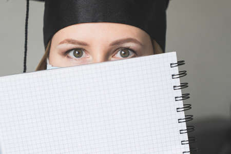 A student in mask is wearing a graduation cape and robe holding blank copy space place concept of the class 2021 coronavirus pandemic of the covid. Graduation student on white backgroundの写真素材