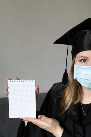 Beautiful blonde woman wearing face mask graduation cap and ceremony robe holding blank banner,...