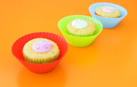 Three vanilla muffins with icing in plastic cups, with yellow fork on grass table against orange wall. の写真素材