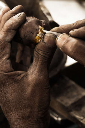 Goldsmith working on an unfinished 22 carat gold ring with big diamond with his hard working hands. Shallow DOF - focus on the ring. Fine gold dust on hands and toolsの写真素材
