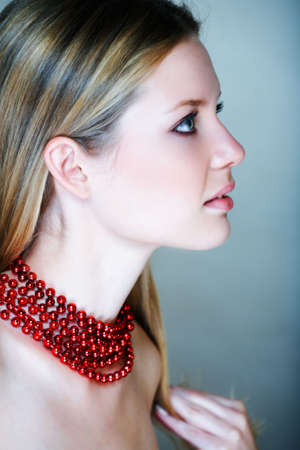 Beautiful young woman with natural make-up and long blonde hair, with red shiny beads in low-key effect on texture background in her early 20sの写真素材