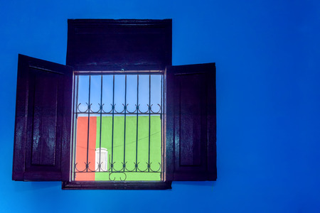 A typical Cuban window with a lattice and a view of the red-green wall with a white window and a blue skyの写真素材