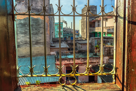 View through an openwork metal lattice on the flaky, dirty-gray stone houses, blue barrels for water, blue sky, smoking labor and houseplantsの写真素材
