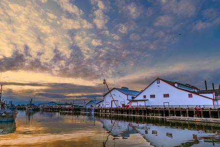 fishing seiners in the harbor at the wooden pier near the warehouse with a winch at sunset of the day with pink clouds and reflection in the water, white buildingの写真素材