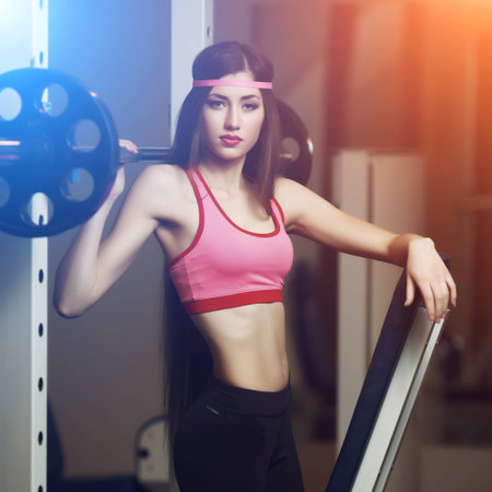 Sexy brunette with very long hair and sporting appearance posing in the gym under the rays of blue and orange light. Young fitness woman in pink t-shirt trainers at the gym.の写真素材