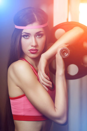 Sexy brunette with very long hair and sporting appearance posing in the gym under the rays of blue and orange light. Young fitness woman in pink t-shirt trainers at the gym.の写真素材