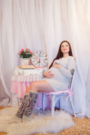 Young Pregnant Brunette, European, stroking her belly sitting on a chair next to the table is a vase of flowers and watchesの写真素材