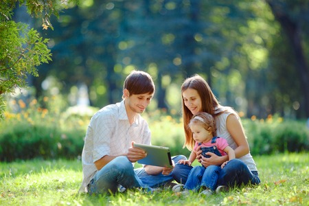 Happy family of three people resting in a city park with a tablet in hands. Father shows in tablet funny pictures. Family sitting on grass and looking at the tablet. Happy family concept of the good life.の写真素材