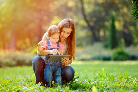 Beautiful mother and kid girl playing at tablet pc outdoors in fall. Happy mother showing her daughter funny pictures your tablet. Happy family, mother and daughter with tablet resting in the park.の写真素材