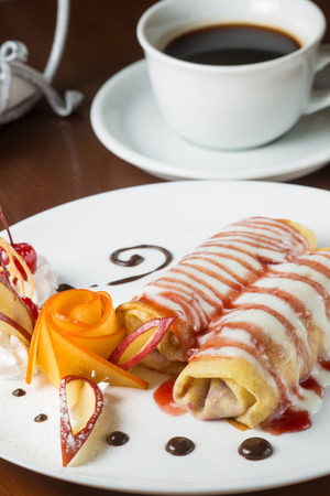 Pancakes with cherry jam beautifully decorated with flowers of fruit and chocolate on a white plate, which stands on a wooden table near the window in the interior cozy cafe. Shallow depth of field.の写真素材