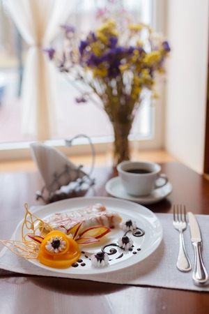 Pancakes with cherry jam beautifully decorated with flowers of fruit and chocolate on a white plate, which stands on a wooden table near the window in the interior cozy cafe. Shallow depth of field.の写真素材