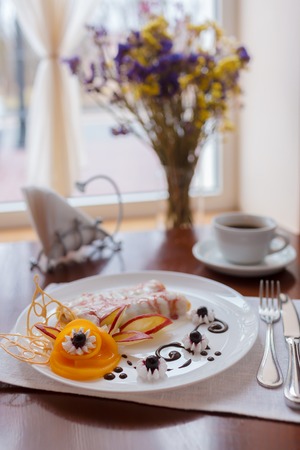 Pancakes with cherry jam beautifully decorated with flowers of fruit and chocolate on a white plate, which stands on a wooden table near the window in the interior cozy cafe. Shallow depth of field.の写真素材