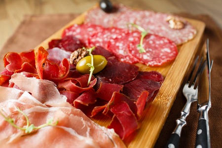 Variety of meats, sausages, salami, ham, olives, laid out on a wooden board close-up, horizontal, are next to a knife and forkの写真素材