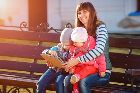 Happy family of three, mother, son and daughter sitting on a bench in the afternoon in the city park and are holding a tablet, a beautiful spring weather, the sun is shiningの写真素材
