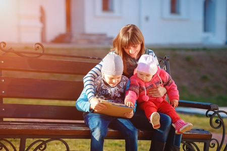 Happy family of three, mother, son and daughter sitting on a bench in the afternoon in the city park and are holding a tablet, a beautiful spring weather, the sun is shiningの写真素材