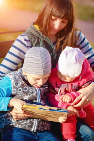Happy family of three, mother, son and daughter sitting on a bench in the afternoon in the city park and are holding a tablet, a beautiful spring weather, the sun is shiningの写真素材