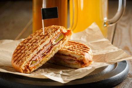 One burger with ham, onion, cucumber and cheese on a wooden plate sprinkled with sesame seeds, paper napkins, small black flag and two glasses of beer on a brown wooden background, close-upの写真素材