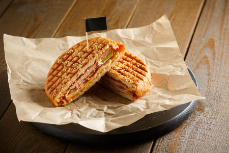 One burger with ham, onion, cucumber and cheese on a wooden plate sprinkled with sesame seeds, paper napkins, small black flag, on a brown wooden background, close-upの写真素材