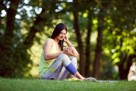 Cheerful athletic woman sitting resting after exercise in the park on the green grass with phone and headphonesの写真素材