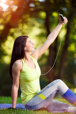 Selfie, Cheerful fitness woman taken pictures of her self phone. Beautiful brunette in sportswear photographs himself in a park in the sunshineの写真素材