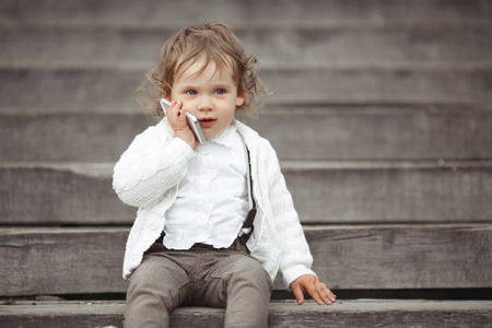 Cute little girl in white knitted sweater talking on mobile phone outdoorsの写真素材