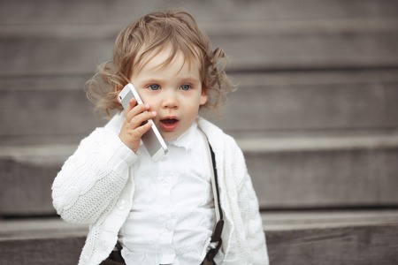 Cute little girl in white knitted sweater talking on mobile phone outdoorsの写真素材