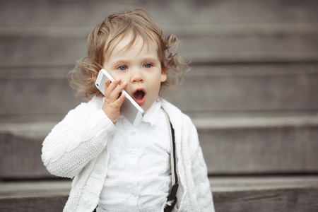 Cute little girl in white knitted sweater talking on mobile phone outdoorsの写真素材