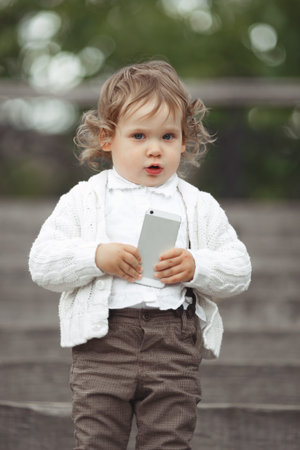 Cute little girl in white knitted sweater playing with mobile phone outdoors
