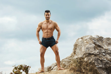 Athletic and muscular man with naked torso stands on top of a mountain. Handsome young guy posing sporty appearance on the sky backgroundの写真素材