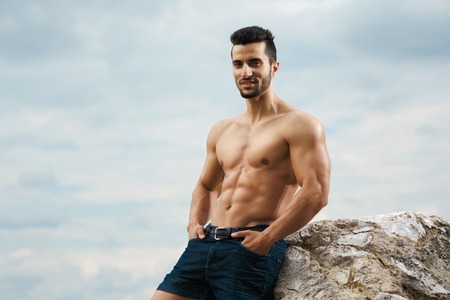 Muscular male torso on a background sky. Athletic and muscular man with naked torso stands on top of a mountain. Handsome young guy posing sporty appearance on the sky backgroundの写真素材