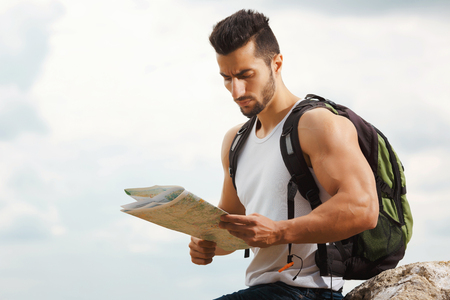 Hiker with a backpack standing on top of the mountain holding a map. Man with map exploring wilderness on trekking adventure. Tourist on the background of the skyの写真素材