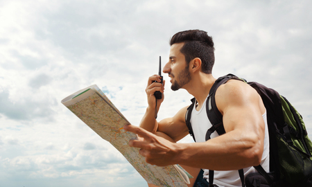 Hiker portrait. Explorer talking via radio station. Young man tourist with a backpack sitting on top of a mountain and holding mapの写真素材