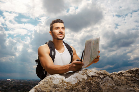 Hiker portrait. Explorer talking via radio station. Young man tourist with a backpack sitting on top of a mountain and holding mapの写真素材